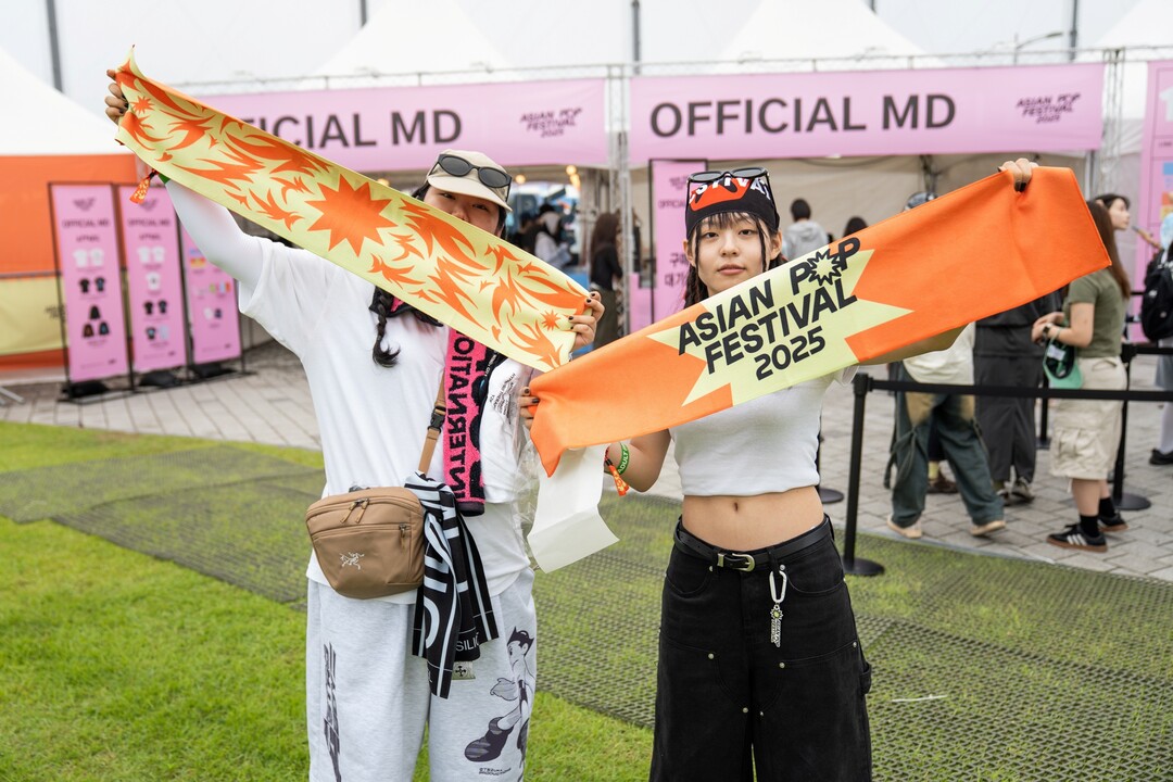 아시안 팝 페스티벌 2025 MD 부스 앞에서 관객들이 공식 슬로건 타월을 들고 포즈를 취하고 있다.